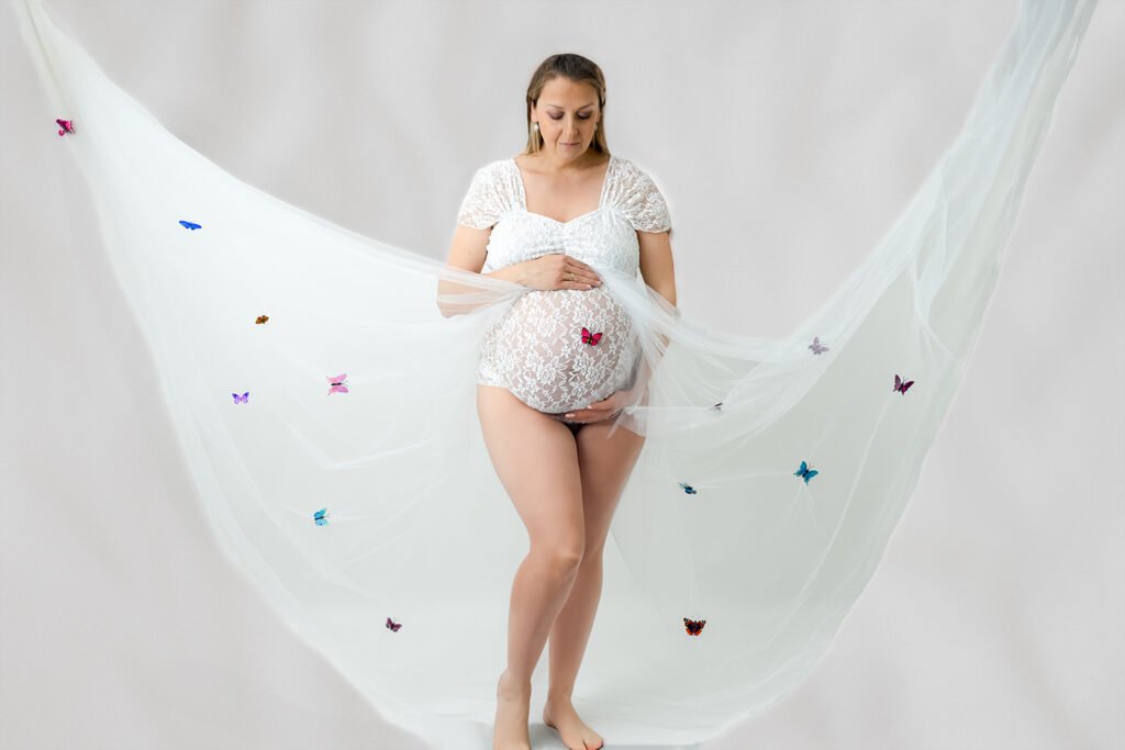 Femme enceinte regardant son vente dans un body dentelle blanc muni d'un voilage sur lesquel sont posés des papillons de plusieurs couleurs.